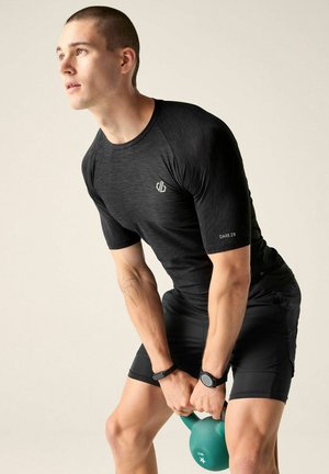 Uomo con abbigliamento sportivo nero che solleva un kettlebell verde, guardando verso l'alto con un'espressione concentrata su uno sfondo semplice.