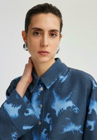 Camicia in denim con un motivo tie-dye blu, colletto classico, chiusura con bottoni e maniche ampie. Tessuto liscio e morbido.