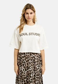 Gekürztes cremefarbenes T-Shirt mit "SOUL STUDIO" im Leopardenmuster, kombiniert mit einem braunen Leopardenmuster-Rock, der eine lockere Silhouette und eine weiche Textur aufweist.