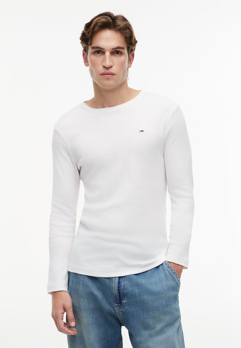 Joven con cabello castaño claro que lleva una camiseta blanca de manga larga con un pequeño logo y vaqueros azules, de pie con una mano en el bolsillo.