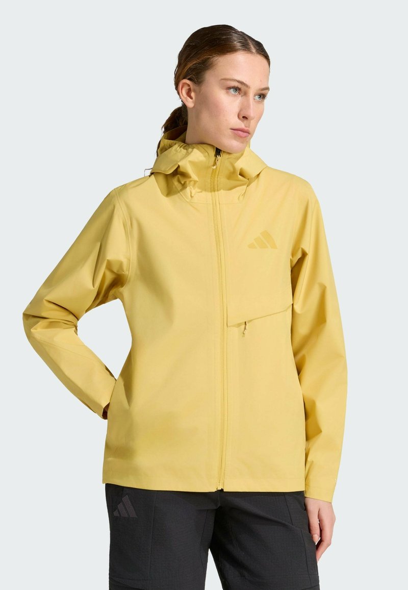 Femme portant une veste de pluie jaune à capuche avec une fermeture éclair devant et un pantalon noir, debout avec une main dans la poche, regardant vers la droite.