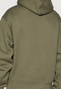Sweat à capuche vert olive avec un tissu texturé, un corps allongé, une capuche réglable avec cordon de serrage, et des poignets et un ourlet côtelés.