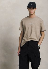 Εκρού βαμβακερό T-shirt με άσπρο κεντημένο λογότυπο, συνδυασμένο με μαύρες cargo παντελόνες με πλαϊνές τσέπες και μαύρο καπέλο, φορώντας ρολόι.