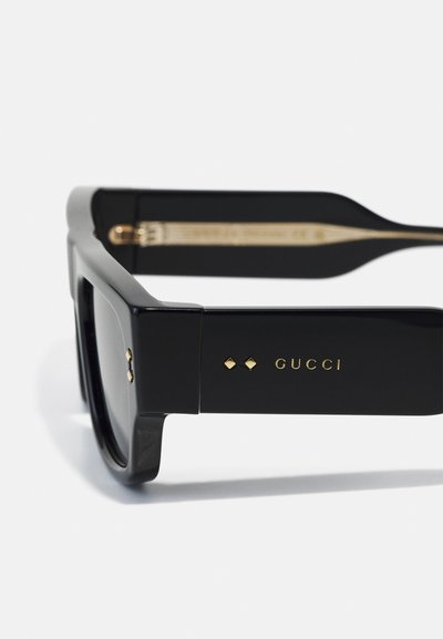 Occhiali da sole neri rettangolari con montatura in plastica lucida. Presentano un logo e dettagli in oro sulle astine. Lenti scure con un design elegante.