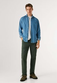 Jasnoniebieska koszula dżinsowa na guziki, noszona na białym t-shircie, zestawiona z ciemnozielonymi spodniami i brązowymi butami na sznurowadła. Model stoi z rękami w kieszeniach.