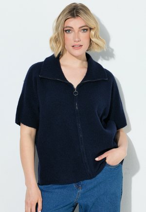 Lyshåret kvinde med kort bølget hår iført en navyblå sweattrøje med kort ærme og lynlås og blå jeans, stående med den ene hånd i lommen.