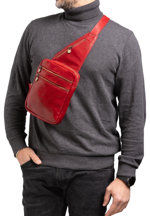 Man draagt een donkergrijze coltrui en zwarte broek met een rode leren schoudertas met meerdere ritssluitingen.