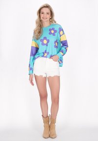 Top de manga larga azul con patrones florales morados y amarillos, combinado con pantalones cortos blancos desgastados y botas de vaquero color marrón.