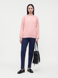 Femme debout portant un sweat-shirt rose, un jean bleu, des mocassins noirs, et tenant un sac cabas en cuir noir sur un fond blanc uni.