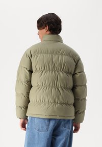 Olivengrønn puffjakke med høy krage, quiltet tekstur og horisontale sømmer. Båret over løse blå denimbukser.
