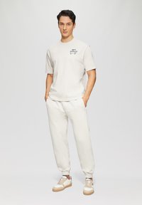 Lichtbeige katoenen t-shirt met korte mouwen en zwarte grafische print, gecombineerd met bijpassende joggingbroek en crèmekleurige sneakers met bruine accenten.
