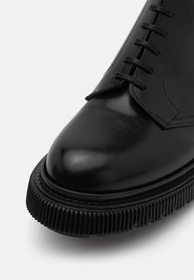 Zwarte leren schoen met een ronde neus, voorzien van een gestructureerde zwarte zool en vlakke zwarte veters, benadrukt door strakke stiksels.