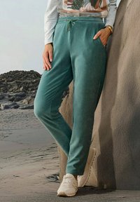 Teal corduroy joggingbroek met een elastische tailleband, zijzakken en een relaxte pasvorm, gecombineerd met witte sneakers op een gestructureerde ondergrond.