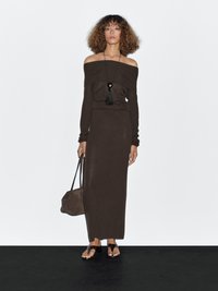 Femme aux cheveux bouclés portant une robe longue à manches longues décolletée sombre, un collier pendentif noir,tenant un sac marron et portant des sandales noires.