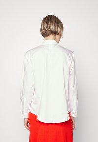 Camicia bianca a maniche lunghe con bottone frontale, orlo curvo e maniche con polsini, abbinata a una gonna rossa brillante a costine. Tessuto liscio.
