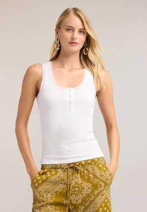 Mujer rubia con pendientes grandes de aro dorados, una camiseta de tirantes blanca acanalada con botones y pantalones con cordón de estampado paisley en color mostaza, con las manos en los bolsillos.