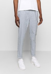 Striped, light blue pants with a tapered fit, featuring a chain detail on the waistband. Worn with a white shirt and white sneakers.