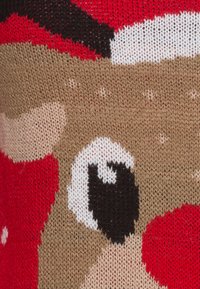 Gros plan sur un tissu tricoté montrant le visage d’un renne avec un grand œil, un nez rouge, une oreille beige, et des détails rouges, blancs et noirs.