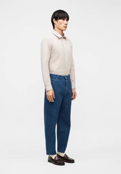 Νεαρός άνδρας όρθιος, φορά μπεζ πουκάμισο με μακριά μανίκια και κουμπιά, μπλε τζιν, ανοιχτόχρωμες κάλτσες και σκούρα καφέ δερμάτινα μοκασίνια, σε λευκό φόντο.