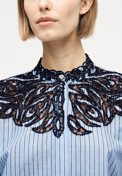 Γυναίκα που φοράει μπλούζα με μπλε και λευκές ρίγες, με μαύρη δαντέλα με λουλουδένιο κέντημα γύρω από τον ψηλό γιακά και τους ώμους.