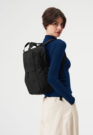 Black backpack made of soft material with a padded back, adjustable straps, and a looped handle, seen from the side against a neutral background.