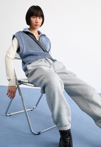Gestreiftes, blau-weißes, ärmelloses Oberteil über einem cremefarbenen Langarmshirt, kombiniert mit grauen Jogginghosen und schwarzen Stiefeln, sitzt auf einem Stuhl.