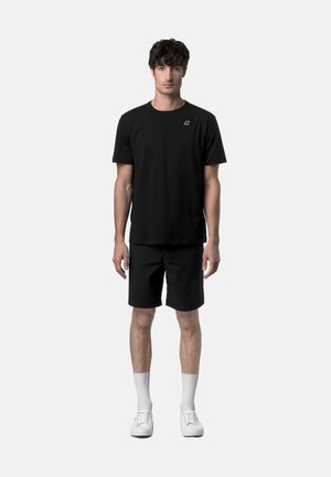 Uomo in piedi che indossa una maglietta nera a maniche corte con logo, pantaloncini neri, calzini bianchi alla caviglia e sneakers bianche, su sfondo neutro.