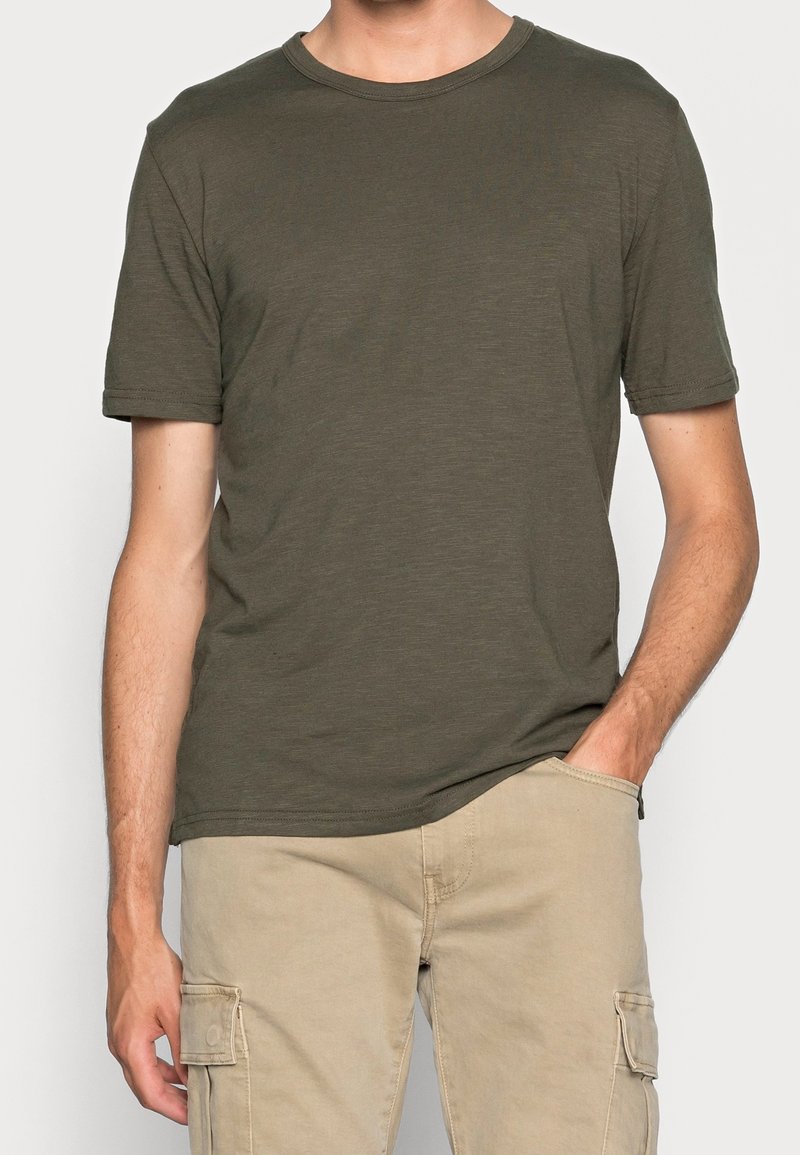 Homme portant un t-shirt à manches courtes col rond de couleur vert olive et un pantalon cargo beige, avec une main dans la poche, sur un fond uni.
