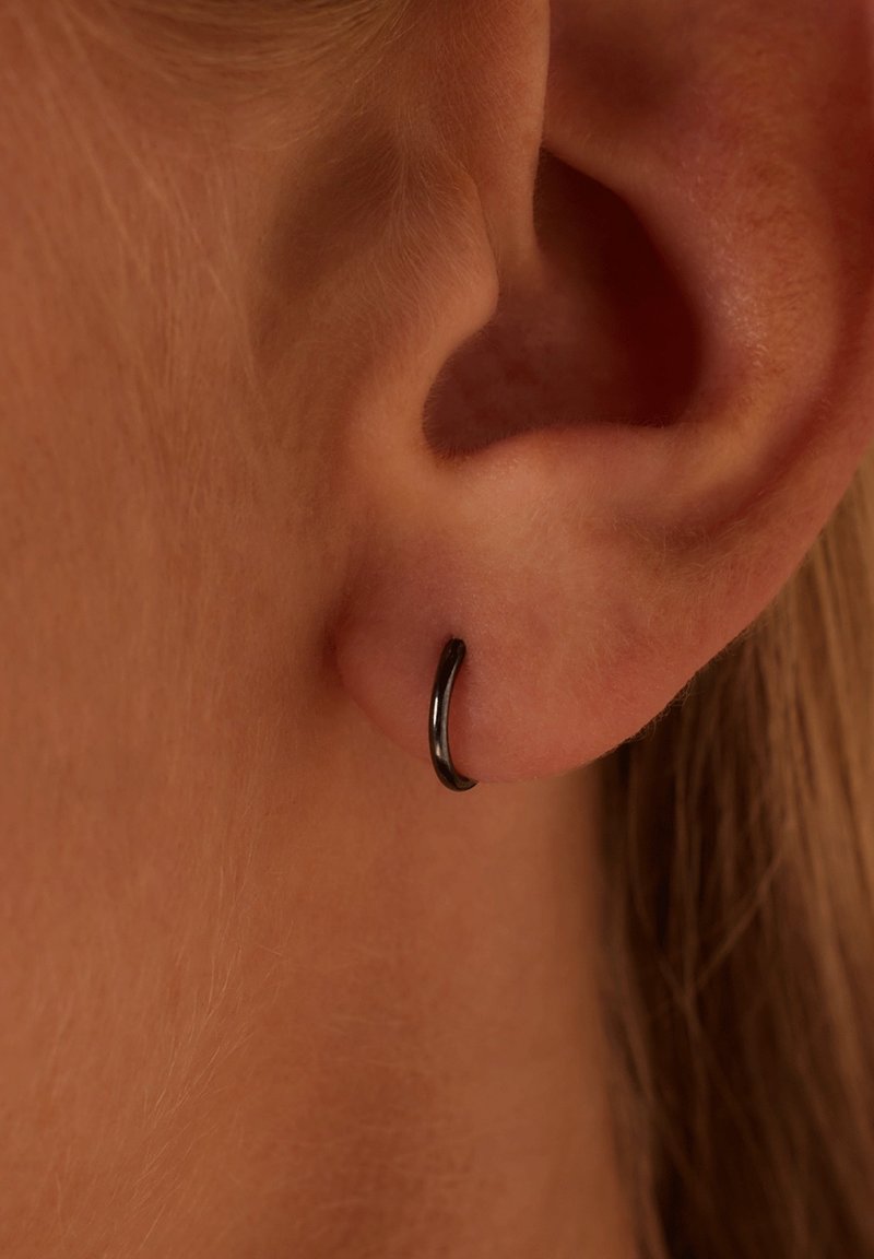 Gros plan sur une oreille gauche à peau claire et cheveux, avec une petite boucle d’oreille noire en anneau dans le piercing du lobe.