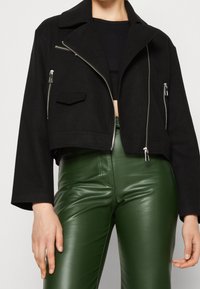 Veste noire cropped avec un col, fermetures éclair argentées et poches avant. Portée avec un pantalon en cuir vert brillant et un haut noir.