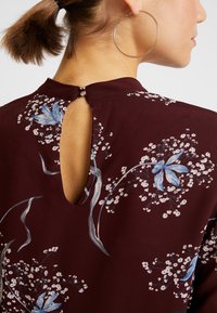 Dos d'une femme portant un chemisier floral bordeaux avec des fleurs bleues et un décolleté en forme de serrure, ses cheveux attachés et une grande boucle d'oreille dorée visible.