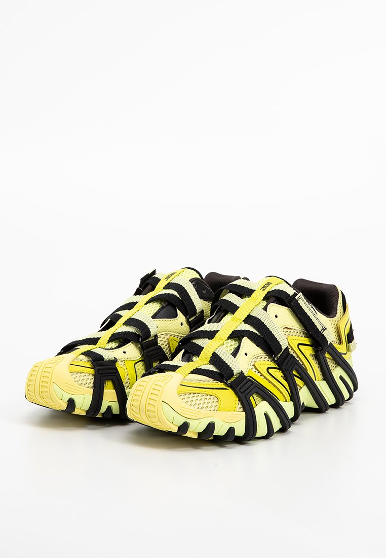 Gelbe Sportschuhe mit einem Mesh-Obermaterial, schwarzen Riemen, strukturierten Sohlen und einem geschwungenen Design mit grünen Akzenten.