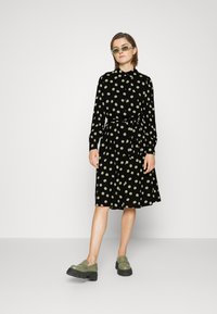 Abito nero con pois verdi, apertura frontale con bottoni, colletto rotondo, maniche lunghe, cintura in vita, lunghezza al ginocchio, abbinato a scarpe verde oliva.
