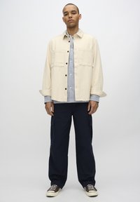 Beige bomulds overshirt med to forlommer, lagd over en blå og hvid stribet skjorte, parret med marineblå cargo bukser og brune sneakers.