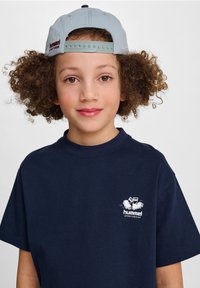 Mörkblå bomullströja med rund hals, med en liten vit logo av tecknade skor på vänster bröst. Ljusblå keps som bärs baklänges.