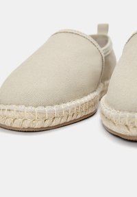 Beige kanvas slip-on sko med vævede jutesåler og en struktureret overflade. Har en lille hælsløjfe for nem påtagning.