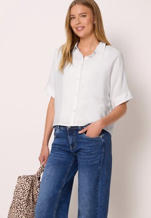 Frau lächelnd, trägt ein locker sitzendes weißes Hemd mit Knöpfen und blaue Jeans, hält eine beige und schwarz gefleckte Tragetasche in einer Hand.