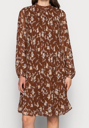 Bruine jurk tot op de knie met lange mouwen, hoge hals en beige en blauwe bloemendessin, gedragen door een staande persoon.