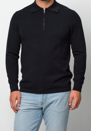 Homme portant un polo en maille noire à manches longues avec une courte fermeture éclair à l'avant et un jean en denim bleu clair, debout devant un fond uni.