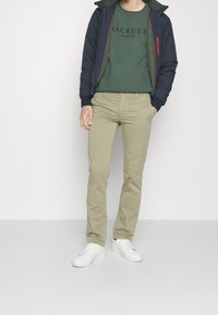 Marinblå bomberjacka, grön sweatshirt med logotyp, khakifärgade byxor och vita sneakers. Jackan har ribbade ärmslut; byxorna har en rak passform.