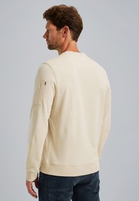 Helles beiges Sweatshirt mit rundem Halsausschnitt und gerippten Ärmelbündchen, ausgestattet mit einer aufgesetzten Tasche am oberen linken Ärmel. Glatte Textur.