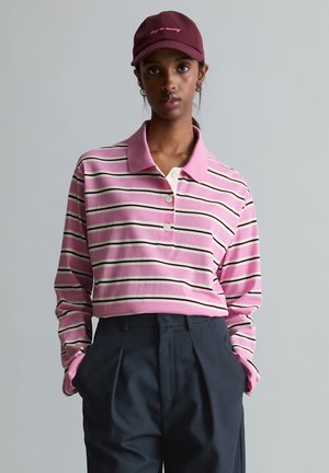 Person mit einer kastanienbraunen Mütze und einem rosa gestreiften Langarm-Poloshirt, das in eine hoch taillierte dunkle Hose gesteckt ist, Hände in den Taschen.