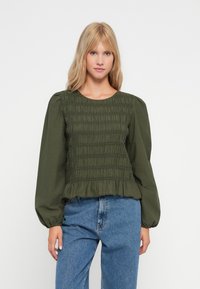 Blouse verte avec smocks, manches bouffantes longues, encolure ronde et détail de ourlet volanté, portée avec un jean en denim bleu clair.