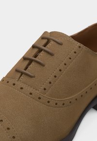 Ανοιχτό καφέ δερμάτινο παπούτσι δετό με λεπτομέρειες μπρογκ και μαύρη σόλα, εμφανίζεται σε κοντινή λήψη.