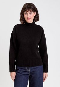 Femme aux cheveux bruns de longueur épaule, portant un pull col roulé noir à côtes et un jean bleu foncé, se tenant devant un fond blanc.
