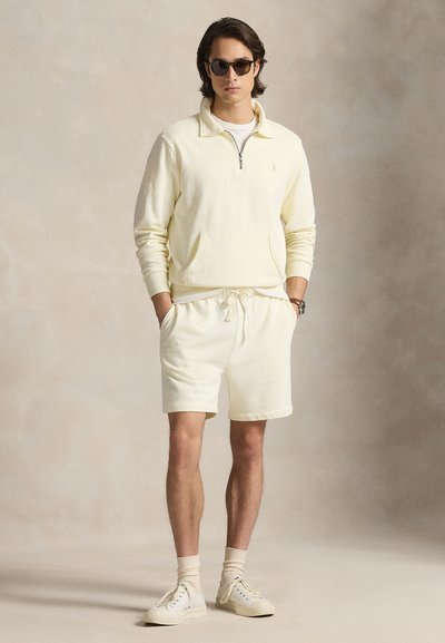 Šviesiai geltonas megztinis su užtrauktuku ir priekiu kišene, derinamas su šviesiai geltonais šortais. Dėvimas su akiniais nuo saulės ir baltais sportbačiais.