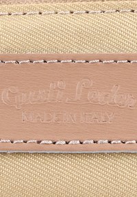 Close-up van beige stof met een gestikt lichtbruin leren label geperst met "Gusti Leder" en "MADE IN ITALY".