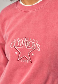 Rosa hettegenser med rund hals. Har hvit brodert tekst "COLORADO COWBOYS" og et stjernedesign med "1998" under. Myk tekstur.