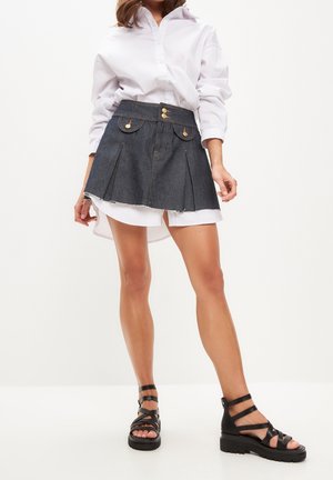 Denim minirok met plooien, gouden knoopaccenten en voorzakken, gecombineerd met een wit oversized overhemd en zwarte sandaaltjes met bandjes.