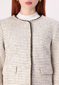Tweed jacket in cream and black with a textured grid pattern, featuring a round neckline, gold-toned buttons, and a contrasting trim.